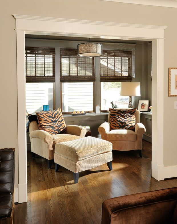 A warm, traditional sitting area viewed through a wide cased opening, furnished with two textured cream armchairs, a matching ottoman, and bold tiger-pattern accent cushions. The room has dark polished timber flooring, a bank of windows with woven blinds, layered white plantation-style shades, classic white decorative trim, and a woven drum pendant light. A leather sofa is partially visible in the foreground of the adjoining room.