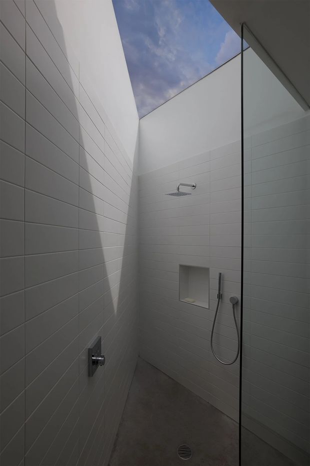 A narrow, minimalist walk-in shower enclosed by a frameless glass screen. The walls are clad in white rectangular tiles with fine grout lines, and the floor is a dark grey concrete-like finish with a linear drain. A recessed wall niche, chrome rain shower, handheld shower, and exposed skylight opening provide the main functional and architectural details.