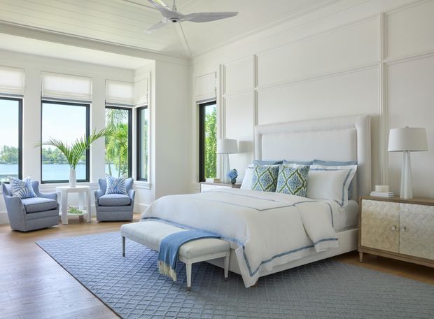 Bright, spacious coastal-style bedroom with a large upholstered bed, blue-and-white bedding, matching armchairs, a bench, bedside tables, and a textured area rug. White paneled walls and ceiling, pale timber flooring, and expansive corner windows provide abundant natural light with a water view beyond.