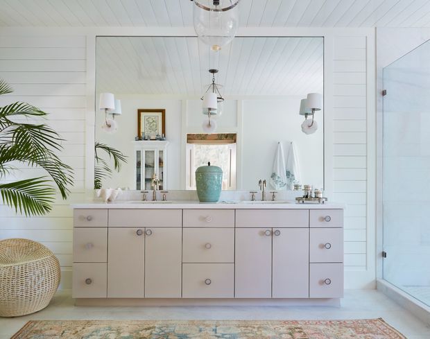 Bright coastal-style bathroom vanity with a long pale built-in cabinet, white countertop and twin basins, framed mirror, wall sconces, glass shower enclosure, decorative turquoise vessel, towels, and tropical greenery. The space features white horizontal wall panelling and a white beadboard ceiling, with a patterned rug and woven basket adding texture.