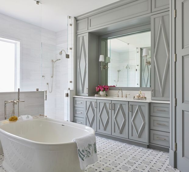 Bright, refined bathroom featuring a freestanding bathtub, glass shower enclosure, double vanity, large mirrored cabinetry, and ornate grey built-in joinery with diamond-patterned fronts. The floor has a geometric grey-and-white patterned tile finish, complemented by pale wall tiles and polished metal fixtures.