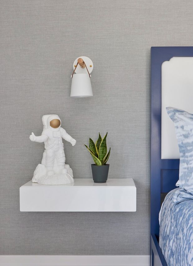 Close-up detail of a styled bedroom wall featuring textured grey wallpaper, a white floating bedside shelf, a small potted snake plant, an astronaut figurine, and a wall-mounted white sconce with brass accents. The edge of a blue-framed bed and blue patterned bedding is visible at right, confirming the bedroom setting.