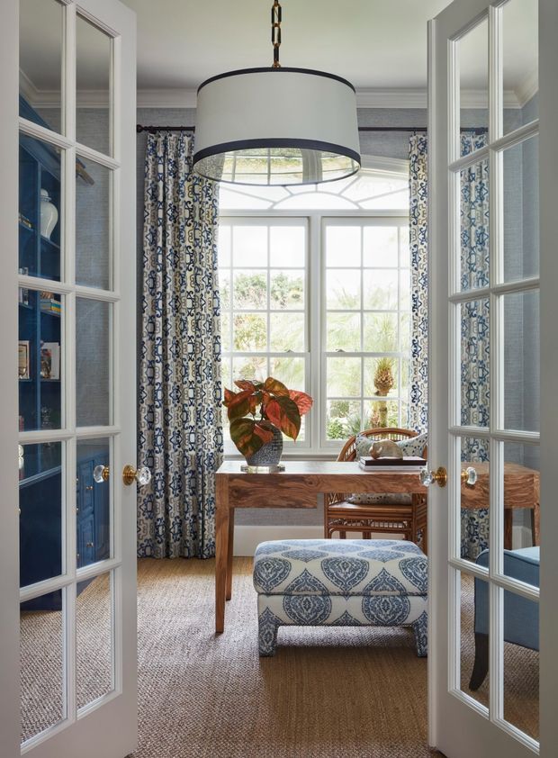 A refined home office viewed through open French doors, featuring a warm wood desk, woven desk chair, patterned upholstered ottoman, blue patterned curtains, blue built-in shelving, and a large multi-pane window. The room has layered traditional detailing, abundant natural light, and a large drum pendant light.