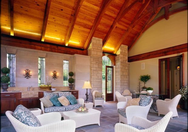 A spacious, elegant living room with a high exposed timber-beam ceiling, warm integrated lighting, pale stone-clad columns and walls, tall narrow windows, dark timber trims, and a large arrangement of white wicker seating around a coffee table. The room opens to an exterior doorway and features traditional decorative furnishings and cabinetry.