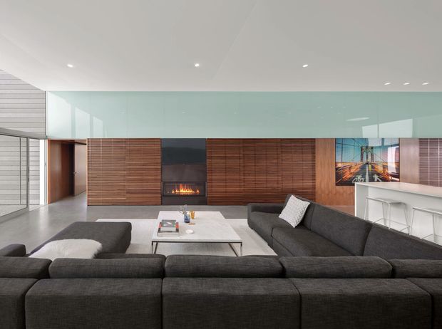 Spacious contemporary living room with a large dark sectional sofa arranged around a pale rectangular coffee table. The room features extensive warm timber wall panelling and built-in joinery, a central black fireplace with visible flame, a pale polished floor, a full-width translucent glass partition or clerestory, recessed ceiling lighting, and a compact white bar or kitchen counter at right. A glazed opening and timber-lined passage are visible at the rear.