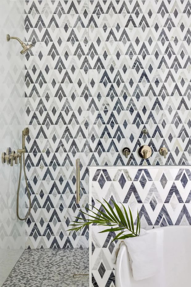 A close-up view of a patterned bathroom featuring a walk-in shower with white, grey and charcoal geometric diamond-patterned wall tiles, matching small-format mosaic floor tiles, and brushed gold shower and tapware. A freestanding white bathtub is partially visible in the foreground with a green palm frond accent.