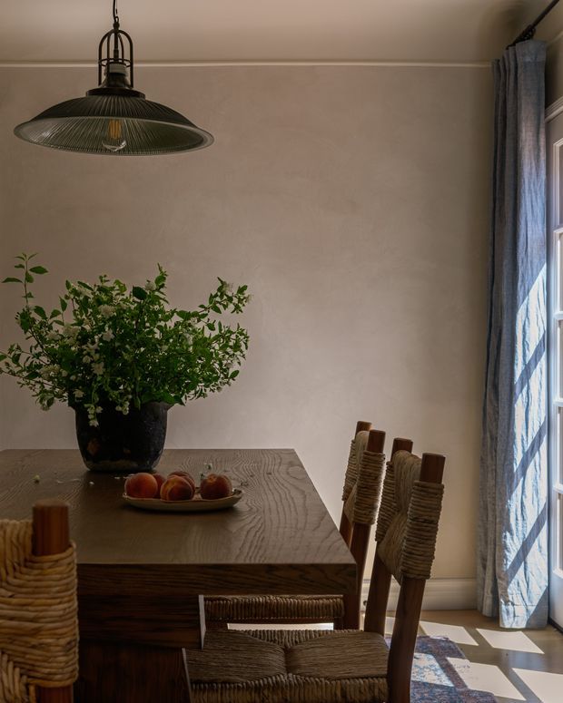 A warm, intimate dining room with a substantial dark wood dining table and woven rustic dining chairs. A large leafy floral arrangement in a dark vessel forms the centrepiece, with apples on a shallow dish. A sculptural dark metal pendant light hangs above, while full-height blue-grey curtains frame a bright multi-pane window. The plastered walls and patterned rug create a refined, natural, softly lit atmosphere.