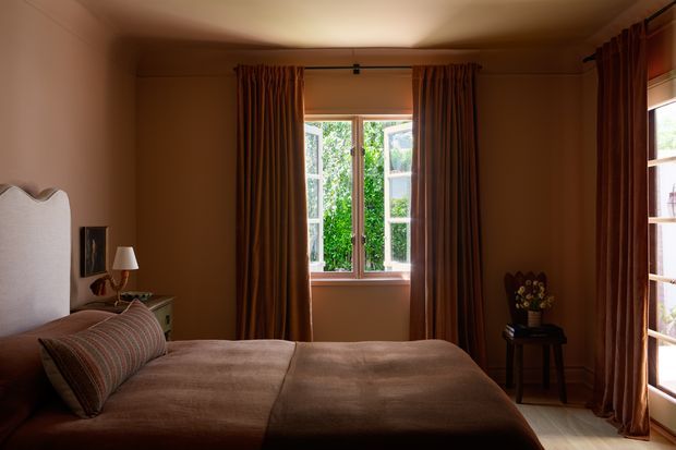 A warm, dimly lit bedroom with a large upholstered bed, scalloped light fabric headboard, layered striped and neutral bedding, and dark rust-brown floor-to-ceiling curtains. Tall timber-framed windows provide views of greenery, while a small bedside table, lamp, artwork, and occasional table with flowers complete the refined traditional setting.