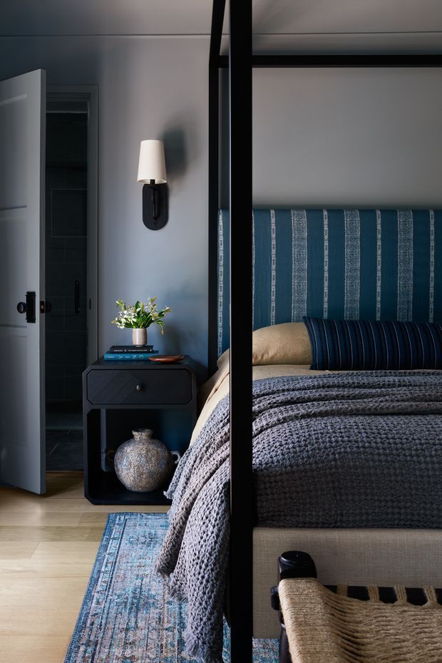 A moody, refined bedroom interior viewed through a black-framed glass partition. The room features a blue upholstered headboard with vertical patterned stripes, layered blue-grey knitted bedding, a dark bedside console, wall-mounted sconce, pale timber flooring, and a patterned blue rug. An open doorway reveals a dark bathroom or dressing area beyond.