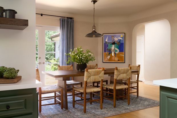 Warm, traditionally detailed dining area viewed between green kitchen cabinetry. A dark timber dining table is surrounded by woven-seat wooden chairs on a patterned rug, with a pendant light overhead, framed artwork, curtains, and an arched opening leading toward another room. Cream walls, timber flooring, and natural textures create a relaxed, refined interior.