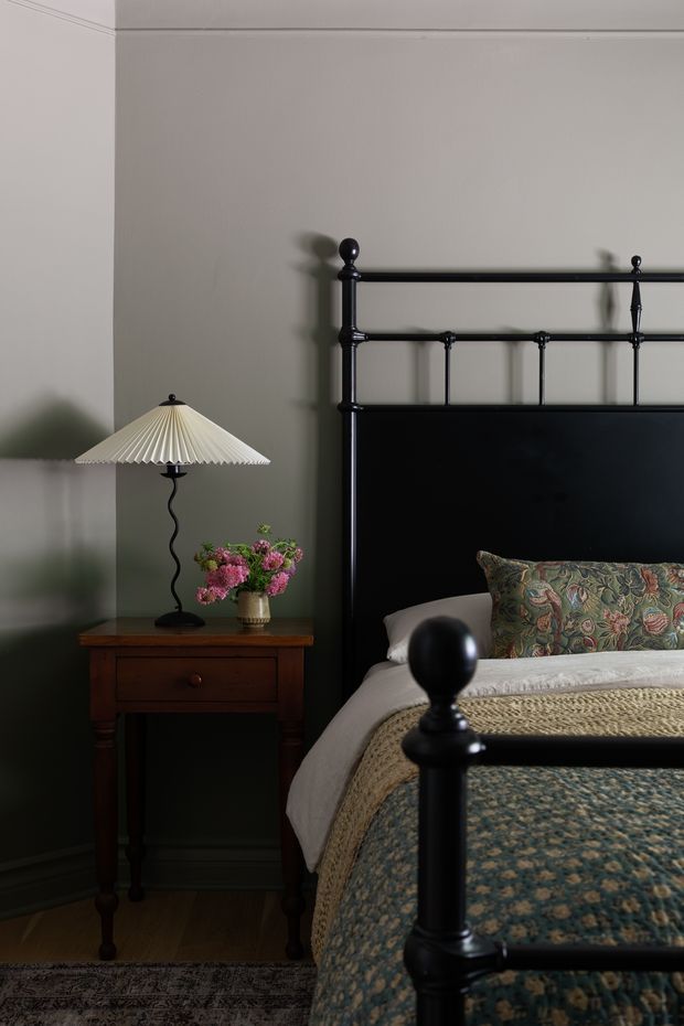 A moody, traditionally styled bedroom vignette featuring a black metal four-poster bed with turned finials, dark patterned bedding, a floral accent cushion, and white bedding. Beside it is a dark timber bedside table with turned legs, a pleated table lamp, and a small vase of pink flowers. The room has muted grey-green walls with decorative ceiling and skirting trims, timber flooring, and a patterned rug.