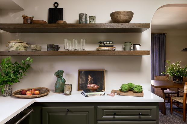 A refined kitchen or dining-room built-in display and serving area with dark green shaker-style cabinetry, a white countertop, and two thick rustic timber open shelves. The shelves hold ceramics, glassware, bowls, books, and decorative objects, while produce and a framed artwork sit on the counter. An arched opening reveals a dining area with timber furniture and dark curtains.