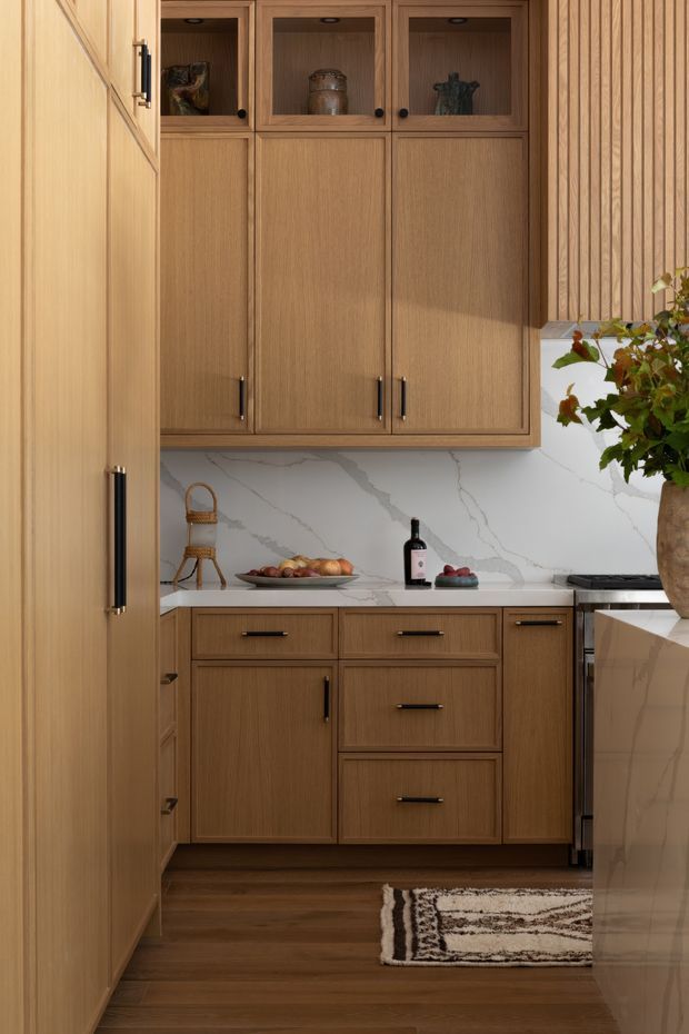 A warm, compact kitchen with floor-to-ceiling natural timber cabinetry, black hardware, a white marble-look stone backsplash and benchtop, and dark timber flooring. The cabinetry includes upper display cupboards with glass fronts, integrated storage, and lower drawers. A partial island or peninsula is visible at right, along with subtle decorative objects and a patterned runner rug.