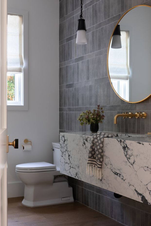 A refined contemporary bathroom featuring a wall-mounted floating vanity with a dramatic white marble-look benchtop and matching fascia, a brass wall-mounted tap, dark grey vertically arranged wall tiles, a round mirror with a thin brass frame, and a modern white toilet. A black-and-white pendant light, roller-shaded window, patterned hand towel, and small floral arrangement add contrast and warmth.