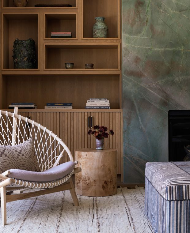 A warm, refined living room featuring a built-in timber shelving and cabinetry wall with display niches, books and decorative objects. A dramatic veined green-grey stone fireplace surround occupies the right side, while a woven white lounge chair, patterned ottoman, cylindrical timber side table, rug and floral arrangement create a relaxed seating area.