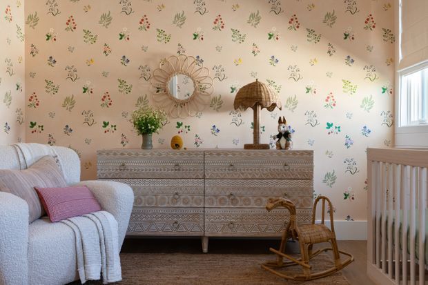 A bright, warmly styled nursery or child's bedroom with whimsical botanical and floral patterned wallpaper, a textured light upholstered armchair, patterned wooden dresser, woven pendant lamp, sunburst mirror, rocking horse, and a wooden cot. The room has a playful vintage-inspired aesthetic with natural textures and soft pastel accents.