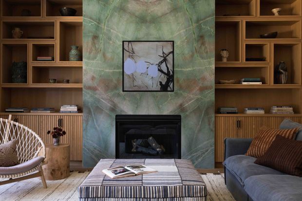 A refined living room centred on a black fireplace set within a dramatic green veined stone feature wall. Warm timber built-in shelving and ribbed cabinetry flank the fireplace, displaying books and pottery. The room includes a blue-grey sofa, woven lounge chair, striped upholstered ottoman, textured cushions, and a pale rug, creating a layered, sophisticated interior.