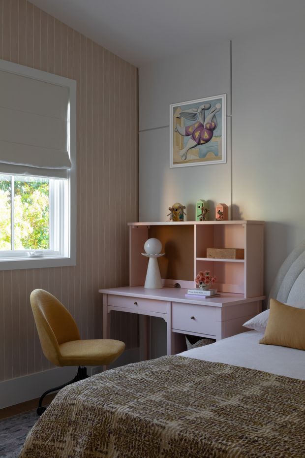 A softly lit bedroom featuring a compact built-in desk and shelving unit, an upholstered mustard desk chair, a partially visible bed with textured brown throw, and a warm grey wall. One wall is clad in vertical pale timber panelling, with a window fitted with a Roman blind and colourful artwork above the desk.