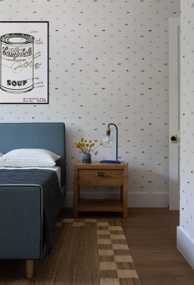 A cozy children's bedroom featuring a blue upholstered bed, white striped bedding, a wooden bedside table with a small blue desk lamp and vase of yellow flowers, playful illustrated wallpaper, framed pop-art artwork, and a brown patterned rug over timber flooring.