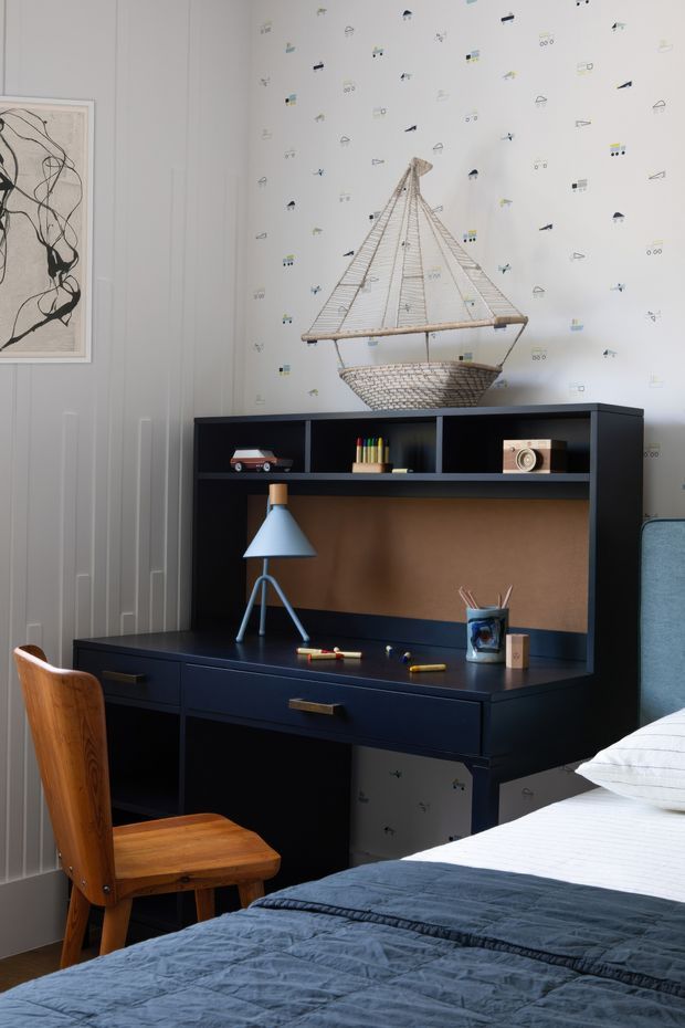 A stylish bedroom with a built-in dark navy or black desk and shelving unit, featuring a warm wood work surface, desk lamp, stationery, books and decorative objects. A mid-century-inspired wooden chair sits at the desk, while the foreground shows a bed with white bedding and a blue-grey quilt. The walls combine vertical panel moulding with playful patterned wallpaper, and framed abstract artwork and a woven hanging boat sculpture add visual interest. The room has a refined, playful, slightly nautical character with a coordinated blue, white and warm wood palette.