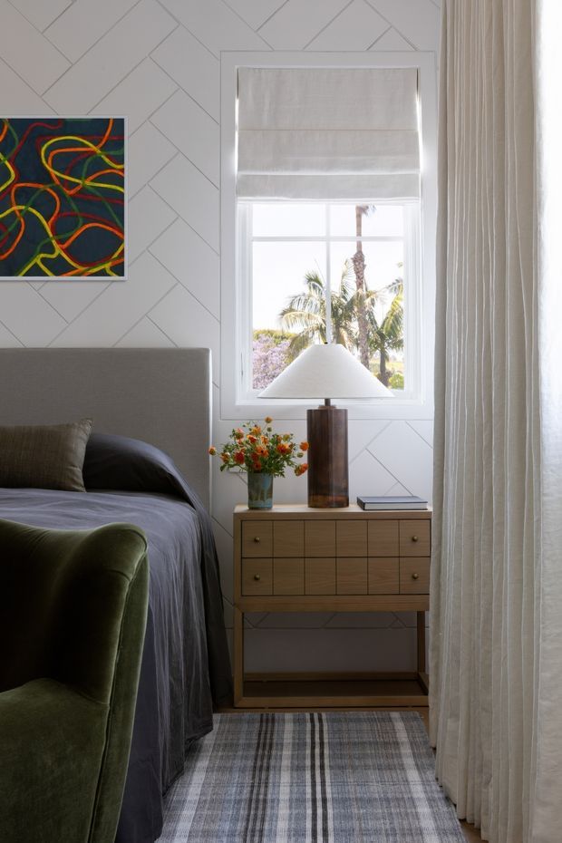 A refined bedroom corner with a grey upholstered bed, dark bedding, and a green velvet armchair beside a mid-century-style wooden bedside cabinet. The white wall features geometric diagonal panel detailing, with a framed abstract artwork above the bed. A window with a Roman blind and full-length textured curtains brings in bright natural light, while a table lamp, flowers, and books accessorise the bedside. A patterned grey rug grounds the space.