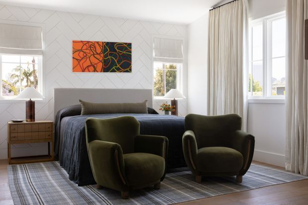 Bright, refined bedroom with a large upholstered bed, two sculptural olive-green armchairs, a timber bedside table, and layered neutral textiles. The room features white geometric wall panelling, tall multi-pane windows, full-height cream curtains, and abstract artwork, creating a polished contemporary-classic atmosphere.