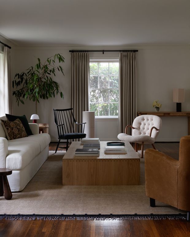 A warm, refined living room with a large white sofa, sculptural upholstered lounge chair, black spindle-back rocking chair, leather armchair, and a substantial timber coffee table. Neutral curtains frame a multi-pane window, while a tall indoor plant, console table, table lamps, patterned cushion, and textured rug add layered detail. Polished dark timber flooring and restrained classic furnishings create a comfortable, sophisticated interior.