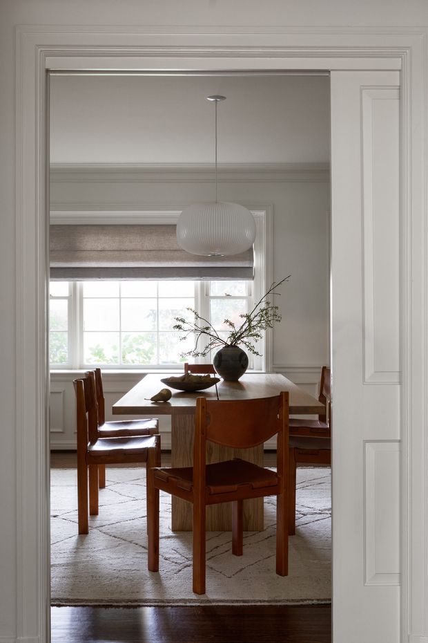 A refined dining room viewed through a wide, heavily moulded doorway. The room features a rectangular timber dining table, four mid-century-style wooden chairs with leather or upholstered backs and seats, a large neutral patterned rug, a sculptural white pendant light, and a dark ceramic vase with branches. Tall multi-pane windows with a woven Roman blind provide soft natural light. The space combines traditional architectural detailing with warm, restrained furnishings.