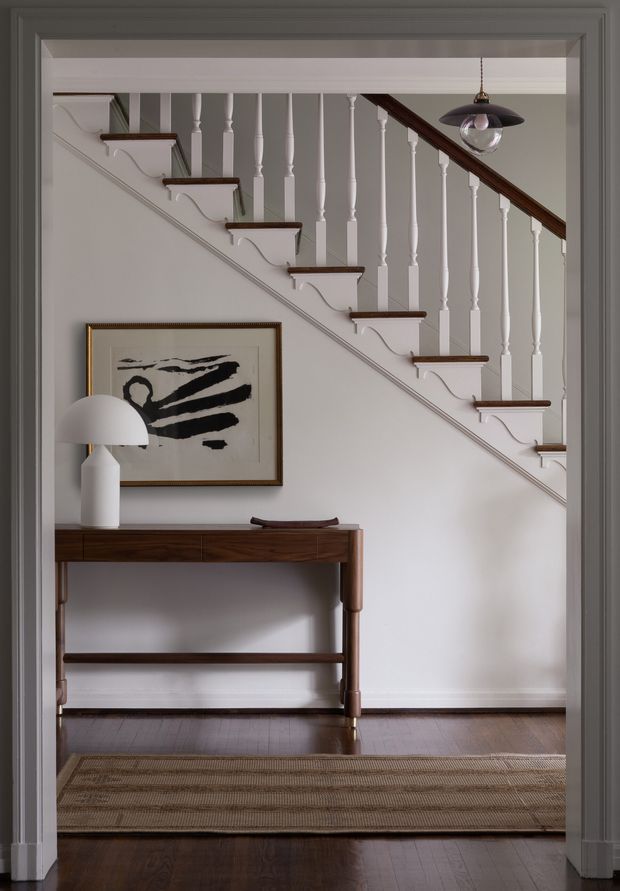 A refined residential hallway or entry passage viewed through a wide doorway, featuring a classic staircase with white turned balusters and dark timber handrail, dark timber flooring, a slim timber console table, framed abstract artwork, a sculptural table lamp, and a pendant light. The restrained palette of white, grey, black, and dark wood creates a calm, elegant transitional interior.