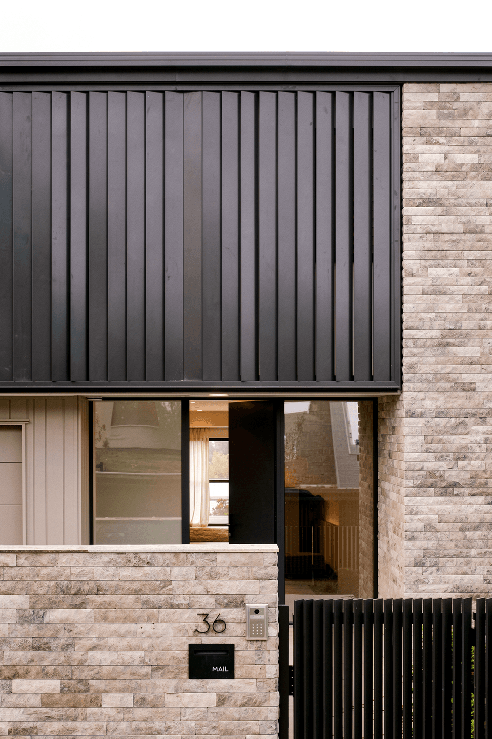Viewed from the front gate, the home presents a refined composition of natural stone and dark architectural detailing. A stone-clad boundary wall and black aluminium slatted gate frame the entrance, while vertical black metal louvres provide privacy and depth to the two-storey street elevation.