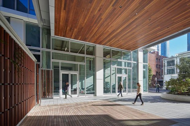 A covered urban pedestrian courtyard or entrance plaza beneath a large warm timber-lined soffit. The space combines extensive floor-to-ceiling glazing and pale structural columns with a vertical timber or terracotta-toned screen wall, broad timber decking, and integrated curved planters. People are walking through the public-facing space, with surrounding city buildings visible beyond.