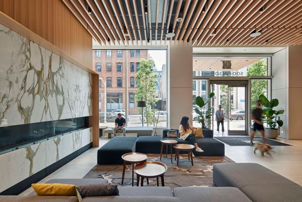 A bright, refined contemporary lobby or communal living area with a large marble feature wall and integrated linear fireplace, warm timber slatted ceiling, full-height glazing, built-in dark upholstered seating, timber coffee tables, and indoor plants. The space opens directly to a glazed street-facing entrance and includes a visible reception or lounge setting.