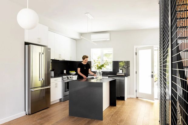 Bright, contemporary open-plan kitchen with white full-height cabinetry, stainless-steel refrigerator and cooking appliances, a large dark island with a pale stone-look benchtop, timber flooring, and a black vertical slatted screen along one side. The room has abundant natural light, a spherical pendant, and glazed doors opening outdoors.