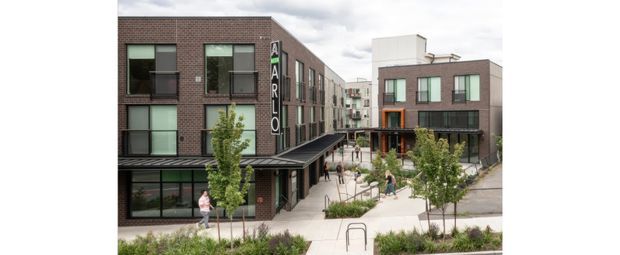 Contemporary multi-storey urban development with dark brick-clad buildings, large rectangular windows, ground-floor commercial or communal spaces, and a landscaped pedestrian courtyard. A covered walkway connects the buildings, with people and young trees indicating an active mixed-use or residential complex.