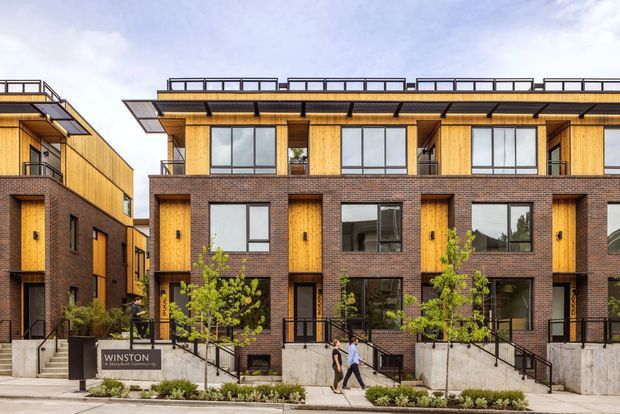 Street-level exterior view of a contemporary multi-storey townhouse development. The buildings combine dark brown brick lower façades with warm vertical timber cladding, large dark-framed windows, projecting timber-clad entry bays, balconies, and broad flat roof overhangs with rooftop guardrails. Concrete retaining walls, steps, black metal railings, young street trees, and a development sign line the frontage.