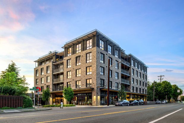 Street-level exterior view of a contemporary multi-storey apartment or mixed-use building on an urban corner site. The building has a prominent brick-clad corner volume, dark-framed windows, projecting balconies, recessed ground-floor commercial glazing, and rooftop terraces. Mature landscaping, adjoining low-rise buildings, utility lines, and a wide road establish an urban suburban streetscape.