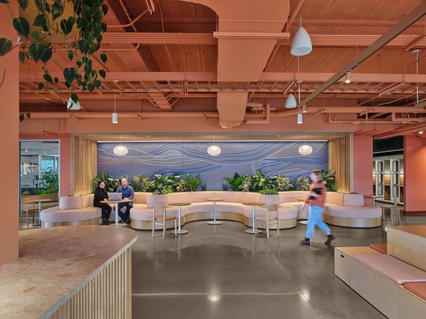 A spacious contemporary commercial lounge or reception area with a warm terracotta-pink palette. Curved cream upholstered banquette seating surrounds small round tables, backed by lush indoor planting and a large blue-and-white wave-pattern feature wall. The exposed ceiling shows painted ductwork, cable trays, pipes, and suspended pendant and track lighting. A stone or concrete-look counter occupies the foreground, while people are present in the seating area and walking through the space.