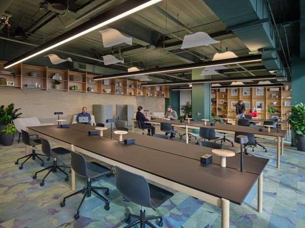 A spacious contemporary open-plan office with long communal worktables, ergonomic task chairs, several seated workers, and extensive open shelving along the walls. The space features exposed services and ductwork, linear LED lighting, sculptural suspended acoustic panels, indoor plants, and colourful patterned carpet tiles. Glazed partitions and a meeting or collaboration area are visible toward the rear.