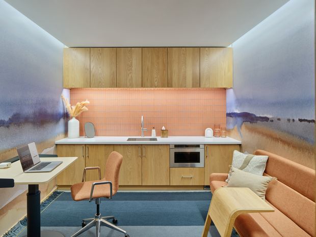 Compact open-plan kitchen and living workspace with warm timber cabinetry, a salmon-pink tiled splashback, integrated oven, white benchtop, and a small dining or work table. Abstract blue-grey and beige wall murals surround the room. A desk with laptop and office chair sits to the left, while a peach upholstered sofa and pale wood side table occupy the right. Recessed ceiling lighting and a dark blue rug create a contemporary, softly colourful interior.