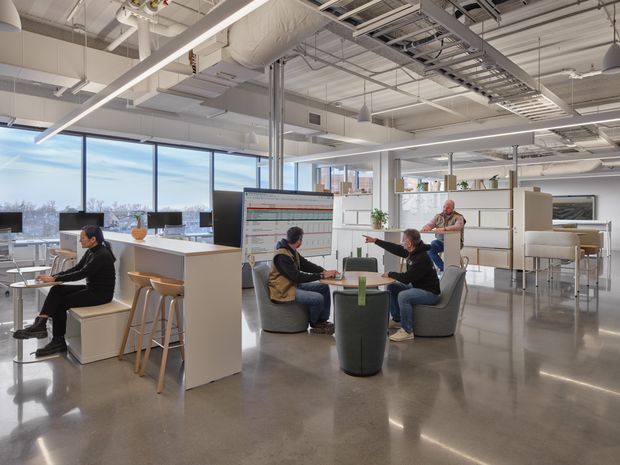 Bright open-plan commercial office with a polished concrete floor, exposed services and ductwork, extensive glazing, white workstations, shelving, and informal seating areas occupied by several people.