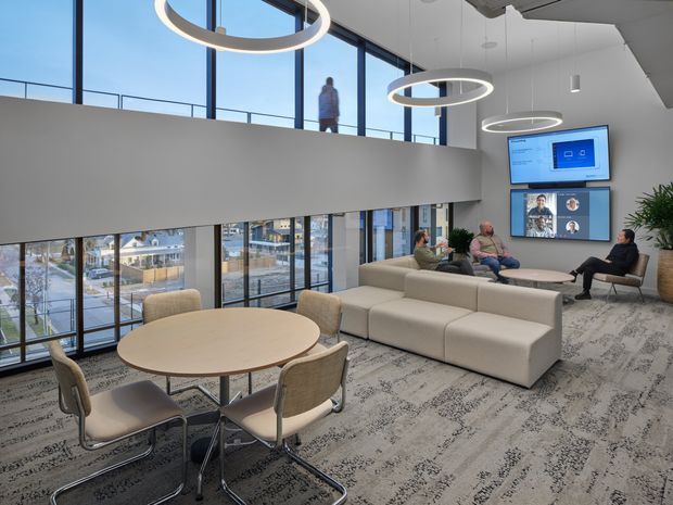 Bright contemporary two-level commercial lounge or workplace breakout area with floor-to-ceiling glazing, a large white sectional sofa, round meeting tables with upholstered chairs, wall-mounted video conferencing screens, ring pendant lights, and patterned carpet. The space overlooks an urban setting and includes a mezzanine-like upper level with a person visible behind a partial-height wall.