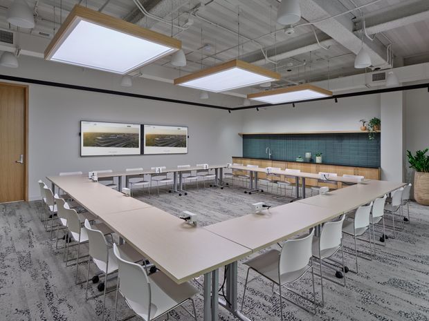 Bright contemporary commercial meeting and training room arranged with U-shaped tables and numerous white chairs. The space features exposed services and ductwork, suspended linear and square pendant lighting, grey patterned carpet, a timber door, a long teal tiled feature wall with a timber counter and open shelving, and wall-mounted display screens.