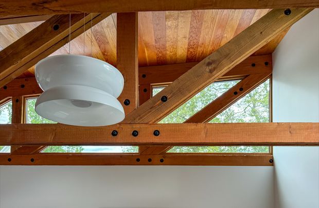 Interior architectural detail showing exposed timber post-and-beam framing with diagonal braces, visible black structural fasteners, a warm timber-lined ceiling, clerestory-style glazing, and a white sculptural pendant light. The view is focused on the upper portion of a room rather than furnishings.