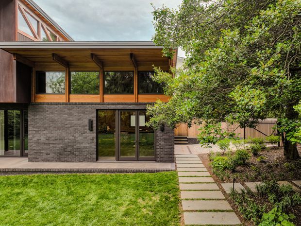 Exterior view of a contemporary multi-level residential building with dark grey brickwork, extensive timber-framed glazing, and a deep timber-clad roof overhang. A paved stepping-stone path leads through a landscaped garden to the glazed entrance, with lawn and mature planting in the foreground.