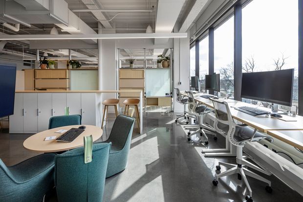 Bright contemporary open-plan office interior with rows of white desks and ergonomic swivel chairs positioned beside large floor-to-ceiling windows. A small round collaborative table with teal upholstered lounge chairs occupies the foreground, while pale wood storage cabinetry, lockers, plants, exposed services, and polished concrete flooring define the workspace. The space has abundant daylight and views of trees outside.