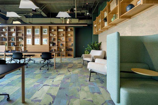 A contemporary open-plan commercial workspace with shared desks, black office chairs, lounge seating, extensive timber shelving and cabinetry, and a high exposed services ceiling. A tall upholstered green acoustic booth occupies the foreground, while patterned carpet tiles in muted green, blue and beige cover the floor. The space combines warm timber joinery with dark painted walls, integrated display storage, and industrial-style ceiling infrastructure.