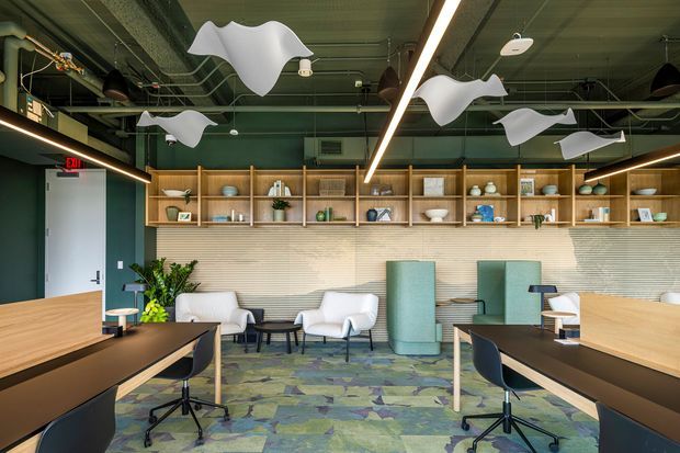 Contemporary commercial office lounge and workspace with exposed services in the ceiling, suspended sculptural acoustic panels, linear LED lighting, open wood display shelving, textured wall finish, upholstered lounge chairs, workstations, and patterned carpet tiles. The space has a refined yet playful, biophilic-inspired atmosphere with muted green, blue, beige, and timber tones.
