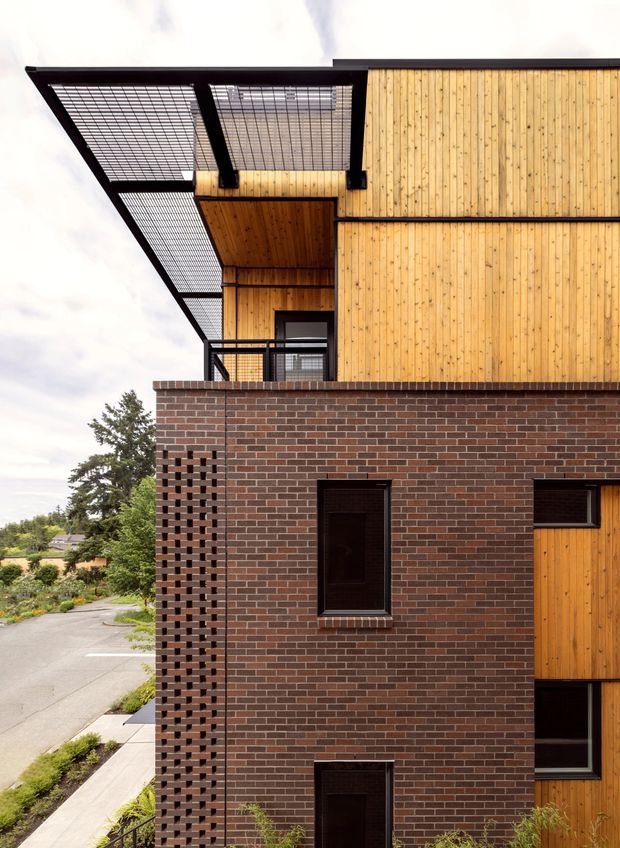 A close-up exterior view of a contemporary multi-storey building combining dark red-brown brickwork with warm vertical timber cladding. The facade features recessed dark-framed windows, a timber-lined upper balcony or recessed outdoor space, and a prominent black steel grid canopy or screened roof structure extending outward. The image also shows a suburban streetscape and landscaping at the edge of the frame.