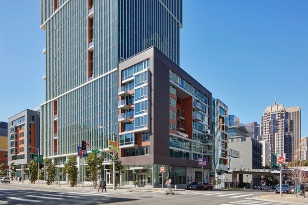 Urban streetscape featuring a contemporary multi-storey mixed-use development with a tall glass curtain-wall tower, adjoining residential or commercial buildings, street-level retail glazing, balconies, mature street trees, and surrounding high-rise buildings. The image is photographed from a broad city intersection under clear daylight.