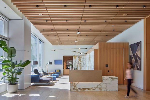 A bright, contemporary commercial lobby or reception area with floor-to-ceiling glazing, a large marble-clad reception desk, warm timber wall panelling, and a slatted timber ceiling with integrated lighting. Blue lounge seating, indoor planting, artwork, and polished stone flooring create a refined hospitality-like entrance.